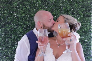 groom and bride kissing in wedding photo booth with boxwood hedge backdrop