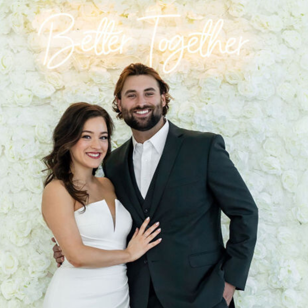 bride and groom in wedding photo booth with flower wall backdrop and better together neon sign