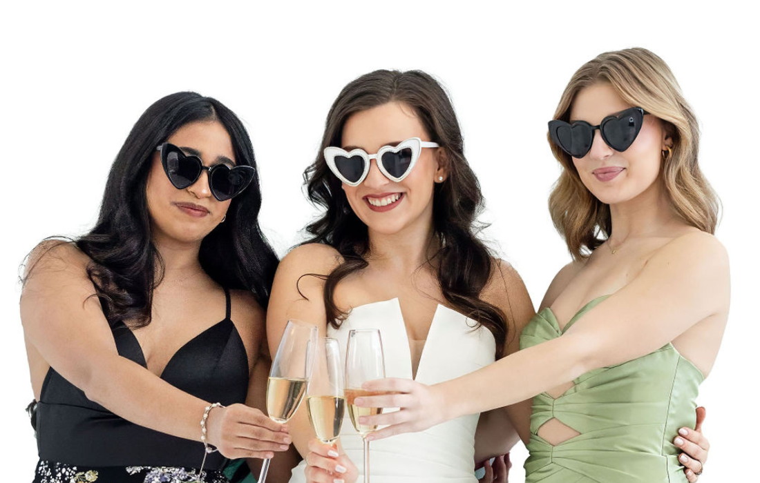 bride with bridemaids posing in wedding photo booth in front of white backdrop toasting champagne glasses