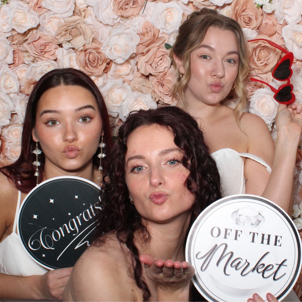 brides-blush-flower2(1000 x 1000 px) three girls modeling wedding dresses posing with engagement prop signs in front of blush flower wall rental in open air photo booth