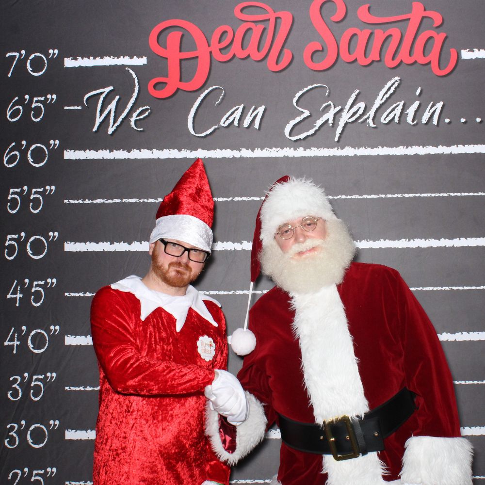 dear-santa-backdrop-elf(1000 x 1000 px) man dressed in red elf suite and a man dressed as Santa standing in front of black backdrop that says dear santa I can explain for holiday photo booth